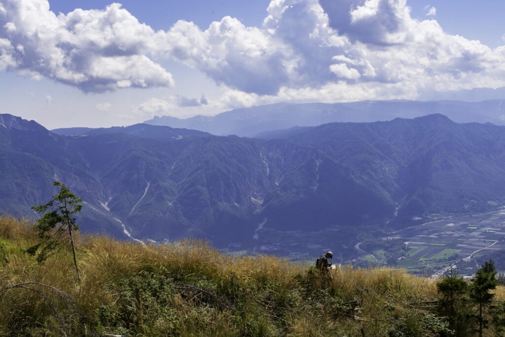 Panoramabild im Vordergrund der mit Gras bewachsene Hang, in dem Dominik Zilg fast versinkt, während er Messpunkte auf seinem Klemmbrett eingibt. Im Hintergrund ein Tal und Bergketten.