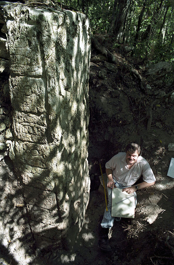Der Altamerikanist Dr. Nicolai Grube steht neben einem Monument der Maya in einem Grabungsschnitt und hat ein Zeichenbrett auf dem Schoß.