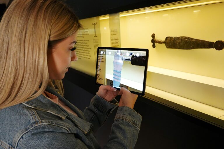 Foto vom Testlauf des AR Spiels Geheimnisgräberei. Zu sehen ist eine Frau vor einer Schauvitrine, in der ein Antennendolch zu sehen ist, Sie hält ein Tablett in der Hand, in dem eine Rekonstruktion des Dolches zu sehen ist.