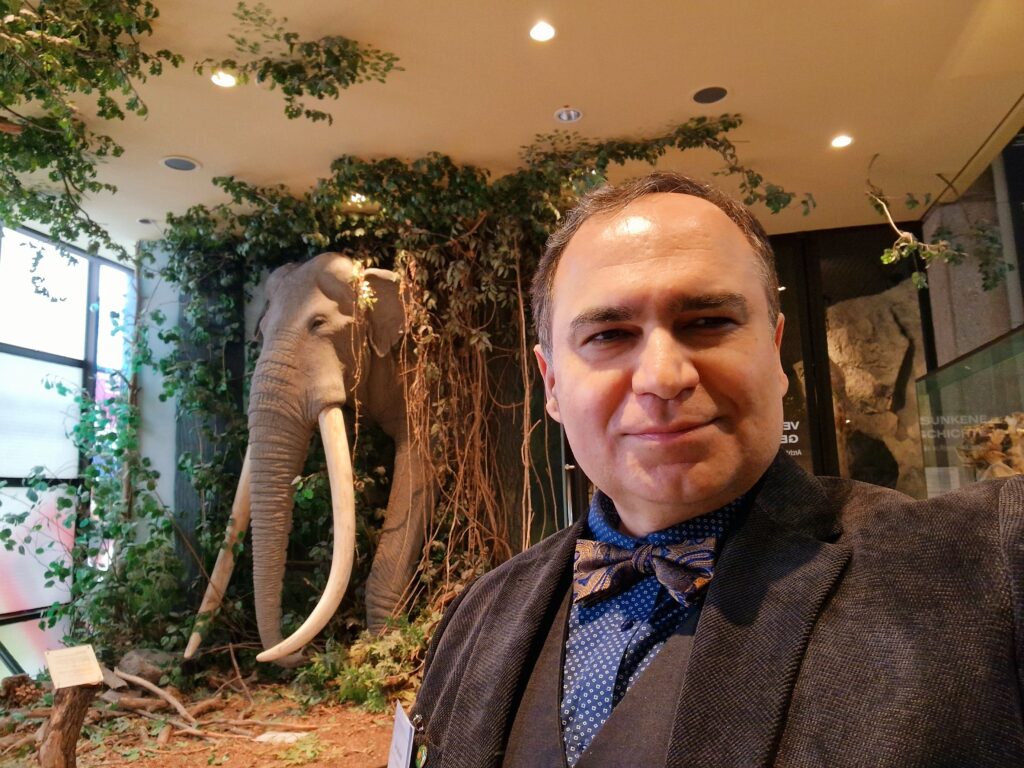 Im Vordergrund ist Nima Nezafati vom DBM zu sehen, der sich selbst fotografiert und im Hintergrund ist ein mit Blättern und Lianen umgebendes Modell eines Elefanten.