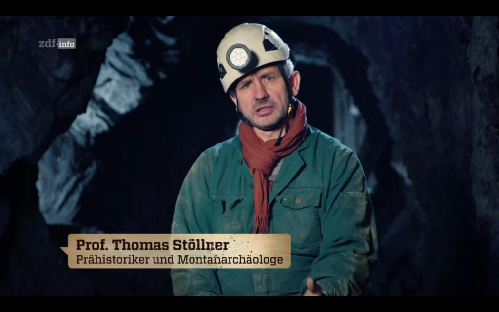 Screenshot aus der Dokumentation Natur macht Geschichte: Erde. Zu sehen ist Thomas Stöllner in Petrolfarbenem Overal, Helm und Grubenlampe, der in einem Stollen sitzt.